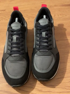 Grå/Svarta sneakers från HUGO - Snygga sneakers från HUGO i grått och svart med röda detaljer på hälen. Skicket på skorna är on top, inga skador eller fläckar.