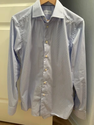 Blårandig skjorta från Eton, s-38 - Klassisk blårandig skjorta från Eton med vit knappstängning och bred krage. Skjortan har lång ärm och är tillverkad i mjuk bomull som känns skön mot huden. Perfekt för dig som gillar stilrena och tidlösa plagg. Passar både till kostym och likväl under en tjocktröja eller ensam med ett par chinos. Observera att skjortan är ”wrinkle free” vilket innebär att den aldrig blir skrynklig och ser ny ut oavsett hur mycket den används. Storlek = S/38