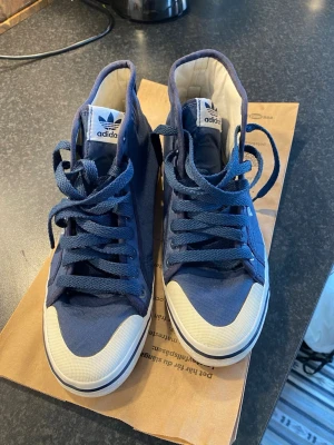 Adidas blå höga sneakers - Snygga blå sneakers från Adidas med högt skaft och klassisk vit tå och sula. Skorna har blå textilsnörning, Adidas-logga på plösen och sidan samt vita detaljer. Perfekta för dig som gillar en sportig och stilren look.