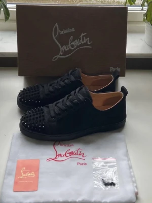 Christian Louboutin svarta sneakers - Svarta sneakers från Christian Louboutin med ikoniska röda sulor och svarta nitar på tån. Skorna har lågt skaft, snörning och är tillverkade i mocka med läderdetaljer. Perfekta för dig som vill sticka ut med en edgy och lyxig stil. Kan även byta mot maison margela !!