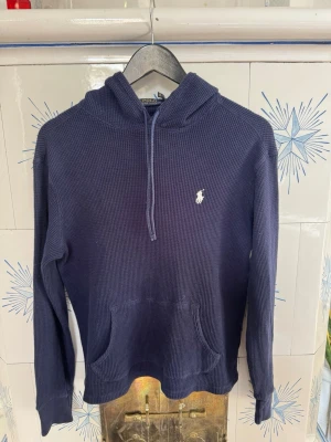Mörkblå hoodie från Polo Ralph Lauren - Mörkblå hoodie från Polo Ralph Lauren i ett grovstickat material med klassisk huva.  Har en vit broderad logga på bröstet och en stor magficka framtill. Perfekt för en avslappnad och stilren look.    Passar både dam och herr 