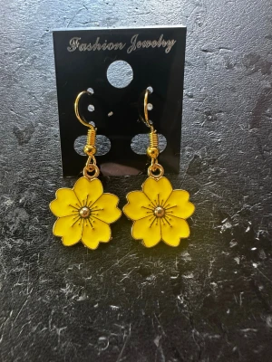 Gula blomformade örhängen - Snygga örhängen i form av blommor med sex kronblad i starkt gul färg och guldfärgade detaljer. Hängena har en metallisk finish och svarta linjer som markerar blombladen. Perfekta för dig som vill sticka ut med färg och lekfull design.