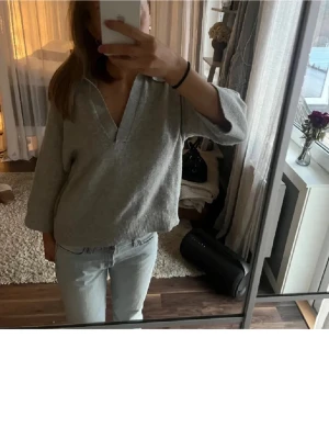 Grå stickad tröja med krage - Mysig grå stickad tröja med bred krage och v-ringning. Tröjan har trekvartsärmar och ribbstickade muddar nedtill. Perfekt för lager på lager och enkel att matcha med jeans eller kjol. Materialet ser ut att vara mjukt och bekvämt.