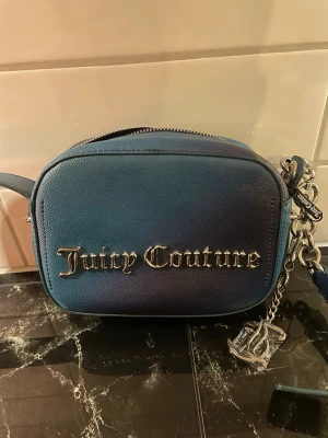 Blå axelremsväska Juicy Couture - Snygg blå axelremsväska från Juicy Couture i läderimitation med silverfärgade detaljer och logga framtill. Väskan har dragkedja, innerfack och dekorativt metallhänge. Perfekt för dig som vill ha en trendig och kompakt handväska.