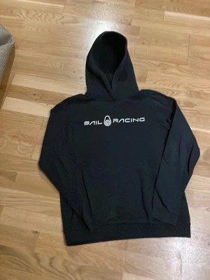 Svart hoodie från Sail Racing - Svart hoodie från Sail Racing med vit logga tryckt över bröstet. Klassisk modell med huva och ribbade muddar vid ärmslut och nederkant. Tillverkad i mjukt material som passar perfekt för en avslappnad stil.