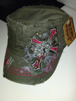 Grön keps med dödskalle och kors - Säljer en grön keps från Legend med råa kanter och slitna detaljer. Kepsen har ett stort broderat kors med dödskalle och nitar på framsidan samt röda och gråa dekorsömmar. Perfekt för dig som gillar en edgy stil.