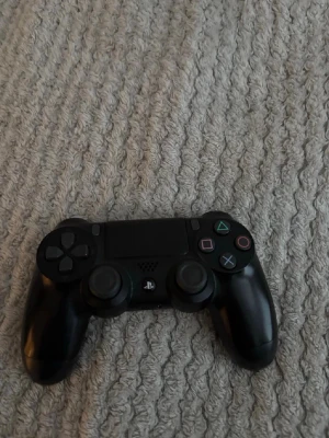 Svart PlayStation 4 DualShock kontroll - Säljer en svart PlayStation 4 DualShock handkontroll från Sony. Den har klassisk design med touchpad, LED-lampa och ikoniska PlayStation-knappar i färg. Tillverkad i plast med greppvänlig form, perfekt för gaming.