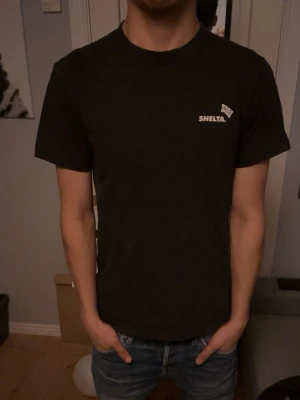 Svart t-shirt från Shelta med tryck - Svart t-shirt från Shelta med liten logga på bröstet och stort grafiskt tryck i vitt på ryggen. Klassisk passform och rund halsringning. Perfekt för dig som gillar streetwear och unika prints.