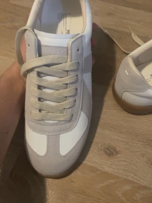 maison margiela  - Stilrena sneakers i vitt och beige med ovandel i mocka och slät textil. Skorna har klassisk låg profil, beige snörning och en snygg brun gummisula. Perfekta för en clean och modern look.