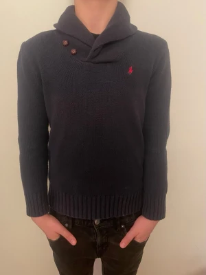  Polo Ralph Lauren stickad tröja - Mörkblå stickad tröja från Polo Ralph Lauren med hög krage och två dekorativa knappar vid axeln. Tröjan har lång ärm, ribbstickade muddar och en liten röd logga broderad på bröstet. Perfekt för dig som gillar klassisk och stilren design.