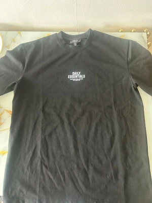 Svart Daily Essentials t-shirt - Säljer en svart t-shirt från Daily Essentials med vit text på framsidan. T-shirten har klassisk passform, rund hals och korta ärmar. Materialet känns som bomull och trycket ger en clean streetwear-vibe.