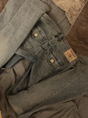 Ljusblå True Religion jeans - Säljer ett par ljusblå jeans från True Religion med ikoniska ficklock och kontrastsömmar på bakfickorna. Jeansen har raka ben och klassisk femficksdesign. Materialet är denim i bomull med en tvättad look. Elr byta till w26-w27