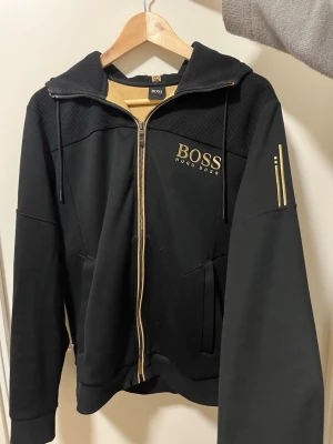 Svart hoodie från Hugo Boss - Snygg svart hoodie från Hugo Boss med dragkedja och guldfärgade detaljer. Stor BOSS-logga på bröstet och huva med dragsko.
