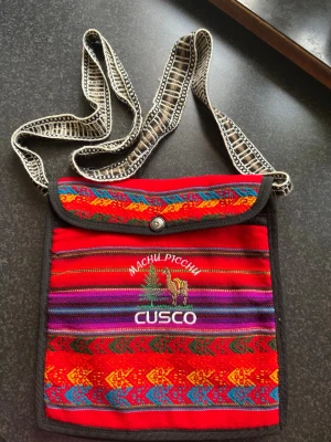 Färgglad axelväska Cusco Machu Picchu - Axelväska i tyg med broderad text 'Cusco' och 'Machu Picchu', samt motiv av lama och berg. Väskan har starka färger som röd, gul, blå och lila i traditionellt mönster. Justerbar axelrem i svartvitt mönster och knappstängning framtill.