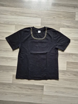 Svart t-shirt med kedjedetalj - Cool svart t-shirt med en chunky kedjedetalj runt halsringningen. Klassisk passform och korta ärmar, perfekt för dig som vill sticka ut lite extra. Materialet känns mjukt och bekvämt, troligen bomull. Enkel men edgy design som passar till det mesta.