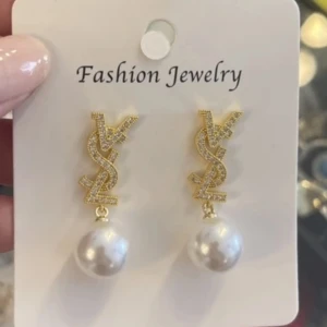 Guldiga YSL örhängen med pärlor - Snygga örhängen i guld med YSL-logga och hängande vita pärlor. Loggan är glittrig och ger en lyxig känsla, medan pärlorna ger en klassisk touch. Perfekta för att lyfta vilken outfit som helst.