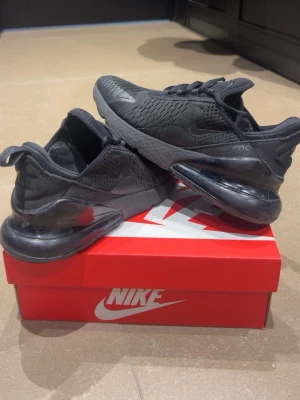 Svarta Nike Air Max 270 sneakers - Nike Air Max 270 sneakers i helsvart med mesh- och syntetmaterial. Skorna har en stor synlig Air-enhet i hälen, snörning och diskreta Nike-loggor på sidan och tungan. Perfekta för dig som gillar sportig och modern stil.