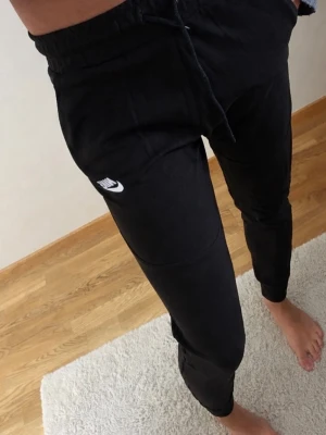 Svarta Nike mjukisbyxor med logga - Säljer ett par svarta mjukisbyxor från Nike med vit logga på vänster lår. Byxorna har dragsko i midjan och muddar vid bensluten för en skön och avslappnad passform. Perfekta för chill dagar eller träning.