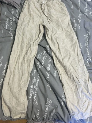 Beige linnebyxor med dragsko - Säljer ett par beige linnebyxor med rak passform och elastisk midja med dragsko. Byxorna är tillverkade i ett tunt och luftigt linnematerial som är perfekt för varma dagar. De har en enkel design utan synliga fickor eller detaljer.