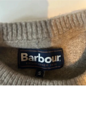 Grå stickad tröja från Barbour - Klassisk grå stickad tröja från Barbour med rund halsringning och ribbade muddar vid ärmslut och nederkant. Tröjan har en diskret Barbour-logga vid nederkanten och är tillverkad i mjukt material som passar perfekt till vardagslooken.
