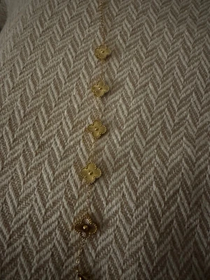 Guldigt armband med blommiga länkar HIMO - Snyggt armband i guldton med flera små blommiga länkar i rad. Kedjan är tunn och elegant, och varje länk har en unik, detaljerad form som ger halsbandet en trendig och stilren känsla. Perfekt accessoar för att lyfta din outfit. Köpt från Himo signature.