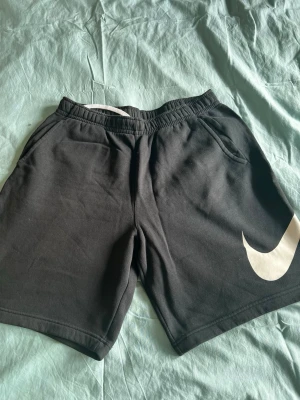 Svarta Nike shorts med stor logga - Säljer ett par svarta shorts från Nike med stor vit swoosh-logga på ena benet. Shortsen har elastisk midja, två sidofickor och en bakficka. Perfekta för chill dagar eller träning. Tillverkade i mjuk bomull för skön känsla.