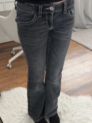 Grå bootcut jeans med broderi - Säljer ett par grå bootcut jeans med snygga kontrastsömmar och broderade bakfickor med blommönster och små nitar. Jeansen har låg midja och klassisk femficksdesign. Perfekta för dig som gillar detaljer och en avslappnad men trendig look.