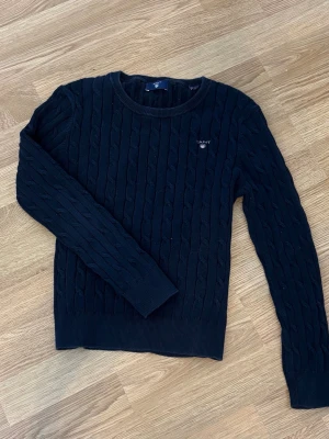 Marinblå kabelstickad tröja Gant - Kabelstickad mörkblå tröja från Gant med rund halsringning och diskret logga broderad på bröstet. Tröjan har långa ärmar och ribbade muddar vid ärmslut och nederkant. Perfekt för lager på lager och klassisk stil.