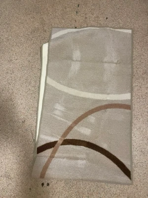 Beige matta med grafiskt mönster - Säljer en rektangulär matta i beige med grafiskt mönster i brunt, ljusbrunt och vitt. Mattan har mjuk yta och diskreta färger som passar bra i de flesta rum. Perfekt för att ge lite extra stil till hemmet.