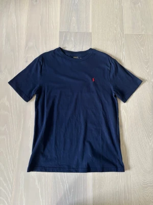 Marinblå t-shirt från Polo Ralph Lauren - Klassisk marinblå t-shirt från Polo Ralph Lauren med liten röd logga broderad på bröstet. Tillverkad i mjuk bomull och har en normal passform med korta ärmar. Perfekt basplagg för en clean och stilren look. Pris går att diskutera. Vid intresse är det bara att höra av sig 