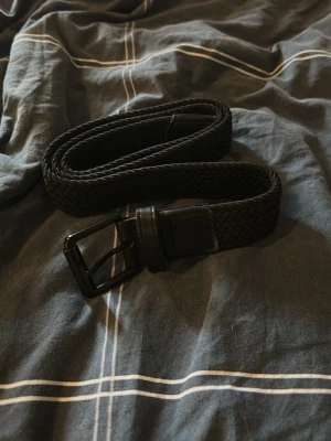 Svart flätat bälte med metallspänne - Snyggt svart flätat bälte från H&M med matt svart metallspänne. Bältet har en stilren design och passar perfekt till jeans eller chinos. Gjort i najs syntetmaterial.