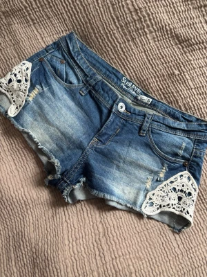 Lågmidjade jeansshorts - Säljer ett par blå lågmidjade  jeansshorts från Sublevel med slitningar och rå kant. Shortsen har vita spetsdetaljer på sidorna och klassisk femficksdesign. Mått finns på bilderna. Perfekta för sommaren och ger en avslappnad men söt look.
