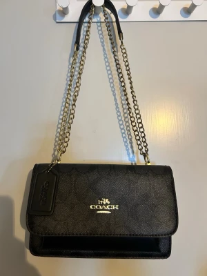 Svart handväska med kedja från Coach - Svart handväska från Coach med guldfärgad kedjerem och logga framtill. Design i läder med diskret mönster och en tag med Coach-logga. 