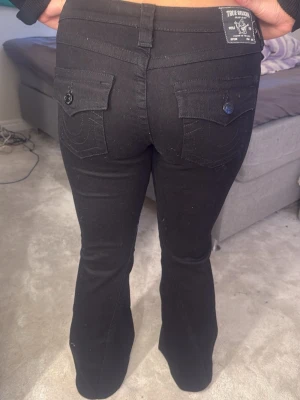 Svarta bootcut jeans True Religion - Säljer ett par svarta bootcut jeans från True Religion med fickor och knappar bak. Jeansen har låg midja, klassiska sömmar och är tillverkade i stretchigt denimtyg. Perfekta för dig som gillar en tidlös och cool look.