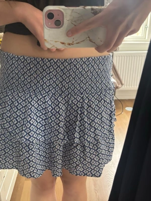 Blåvit mönstrad kortkjol - Kortkjol med blått och vitt geometriskt mönster. Kjolen har en lätt utsvängd form och volang nedtill för en flowy känsla. Perfekt för varma dagar och enkel att matcha med olika toppar.