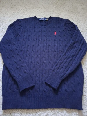 Mörkblå kabelstickad tröja Polo Ralph Lauren - Kabelstickad mörkblå tröja från Polo Ralph Lauren med rund halsringning och klassisk röd broderad logga på bröstet. Tröjan har långa ärmar och ribbade muddar vid ärmslut och nederkant. Perfekt för dig som gillar stilren och tidlös design. Passar mer som S.