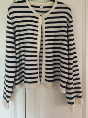 Randig kofta från Gina Tricot - Snygg kofta från Gina Tricot med marinblå och vita horisontella ränder. Koftan har rund halsringning, en dekorativ knapp upptill och ribbade muddar. Perfekt för en avslappnad men ändå stilren look.