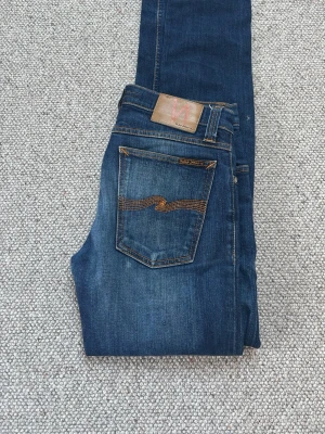 Blå jeans från Nudie Jeans -  Nu Säljer ett par klassiska blå jeans från Nudie Jeans med snygg slitning och orange söm på bakfickan. Modellen har smal passform och är tillverkad i denim med stretch. Perfekta för en avslappnad och trendig look.