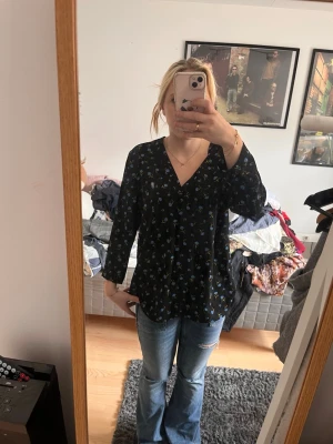 Svart blommig blus med V-ringning - Svart blus med små blommönster i blått och grönt. Blusen har V-ringning och trekvartsärm, vilket ger en avslappnad men ändå stilren look. Passar perfekt till jeans eller kjol för en trendig outfit.