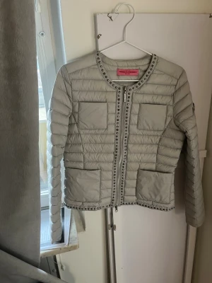 Ljusgrå pufferjacka  - Trendig ljusgrå pufferjacka med rund halsringning och dekorativa nitar längs kanterna. Jackan har fyra stora fickor framtill och quiltad design med horisontella sömmar. Perfekt för dig som vill ha en unik och snygg jacka till hösten eller vintern. Liten sminkfläck på axeln som säkert går bort med sminkborttagning eller i tvätten😊