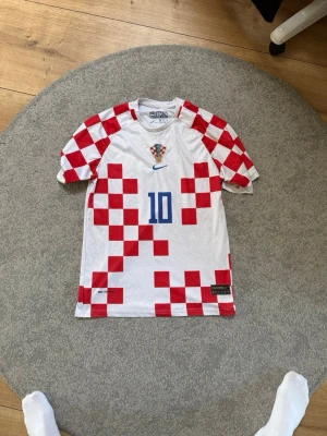 Kroatien fotbollströja Modrić #10 Nike - Säljer en officiell Kroatien fotbollströja från Nike med Modrić och nummer 10 på ryggen. Tröjan är vit med röda schackrutor, blå detaljer och Kroatiens landslagsemblem på bröstet. Perfekt för match eller träning.