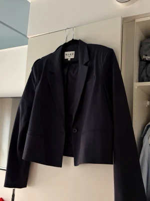 Mörkblå kavaj från NA-KD - Snygg mörkblå kavaj från NA-KD med klassisk krage och en knapp framtill. Modellen är croppad och har långa ärmar. Fint skick!
