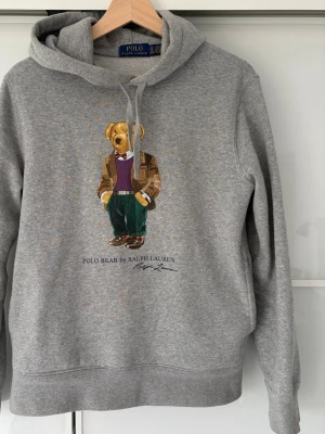 Grå hoodie Polo Bear Ralph Lauren - Grå hoodie från Polo Ralph Lauren med den ikoniska Polo Bear-trycket på bröstet. Klassisk passform med huva och dragsko. Tillverkad i mjukt bomullsmaterial för skön känsla. Perfekt för en avslappnad och trendig look.