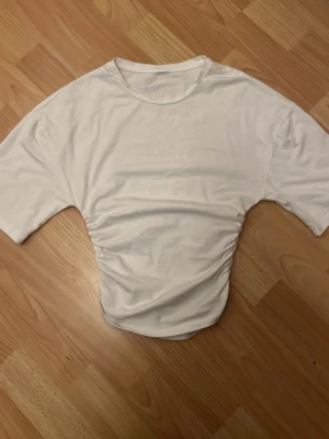 Vit croppad topp med snörning - Trendig vit croppad topp med snörning i sidan och korta ärmar. Toppen har en enkel, stilren design och är perfekt för att matcha med höga jeans eller kjol. Tillverkad i mjukt bomullsmaterial för en bekväm känsla. 