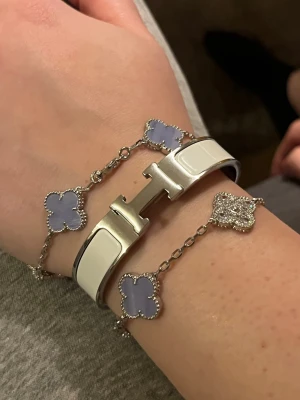 Herme Vitt armband i metall - Snyggt vitt Herme-armband i metall med den ikoniska H-detaljen framtill. Kombinerat med ett kedjearmband med ljuslila fyrklöverformade hängen och gnistrande detaljer. 