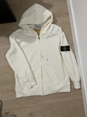 Vit hoodie från Stone Island - Snygg vit hoodie från Stone Island med dragkedja framtill och klassisk svart patch med gult logomärke på ärmen. Hoodien har känguruficka och ribbade muddar. Perfekt för en clean och stilren look.