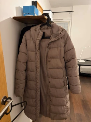 Lång beige dunjacka med huva - Säljer en lång, beige dunjacka med huva och dragkedja samt tryckknappar framtill. Jackan har en quiltad design och är perfekt för kalla vinterdagar. Passar dig som vill ha en stilren och varm jacka med enkel look.