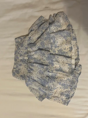 Blommig volangkjol i blått och vitt - Supersöt kort kjol med blått blommönster på vit botten. Kjolen har bred smock i midjan och volanger. Säljs då den inte längre kommer till användning