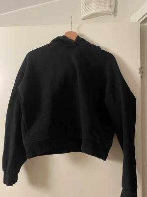 Svart hoodie med huva från Berhska - En svart hoodie med huva och långa ärmar. Hoodien har en avslappnad passform och ribbade muddar vid ärmslut och nederkant. Materialet ser ut att vara mjukt och bekvämt, perfekt för en chill look. Diskret tryck på ryggen.