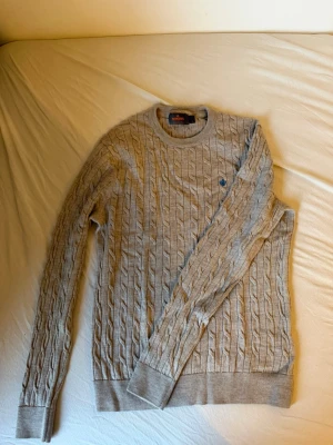 Beige kabelstickad tröja Morris - Snygg beige kabelstickad tröja från Morris i 100% merinoull. Tröjan har rund halsringning, ribbade muddar och en liten blå logga broderad på bröstet. Perfekt för dig som gillar klassisk och stilren design.